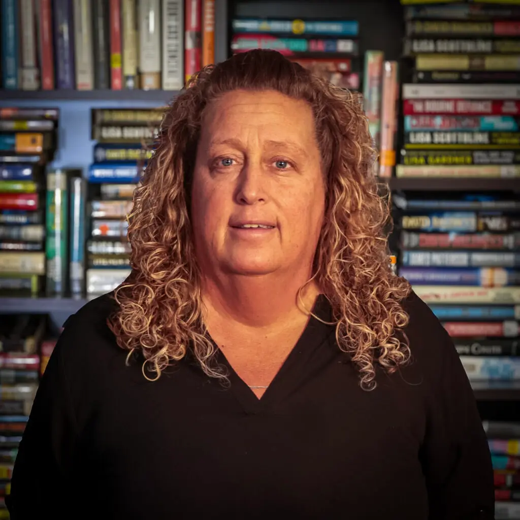 Close-up portrait of a woman with curly blond hair wearing a black top, standing in front of a bookshelf filled with colorful books.