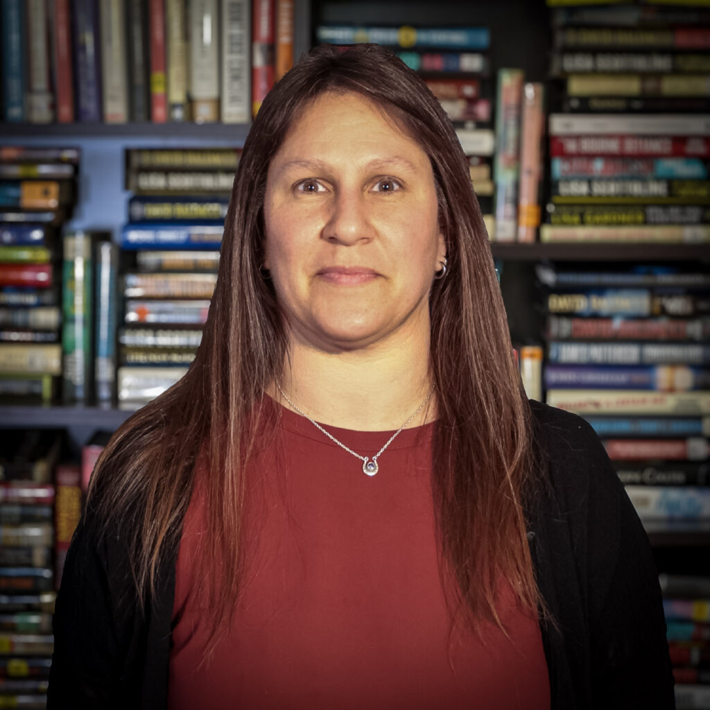 Close-up portrait of a woman with brown hair wearing a red top and black cardigan, standing in front of a bookshelf filled with books.