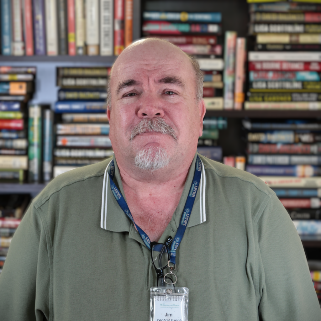 Close-up of an older man with a grey goatee wearing a blue lanyard and badge, standing in front of a bookshelf filled with books.