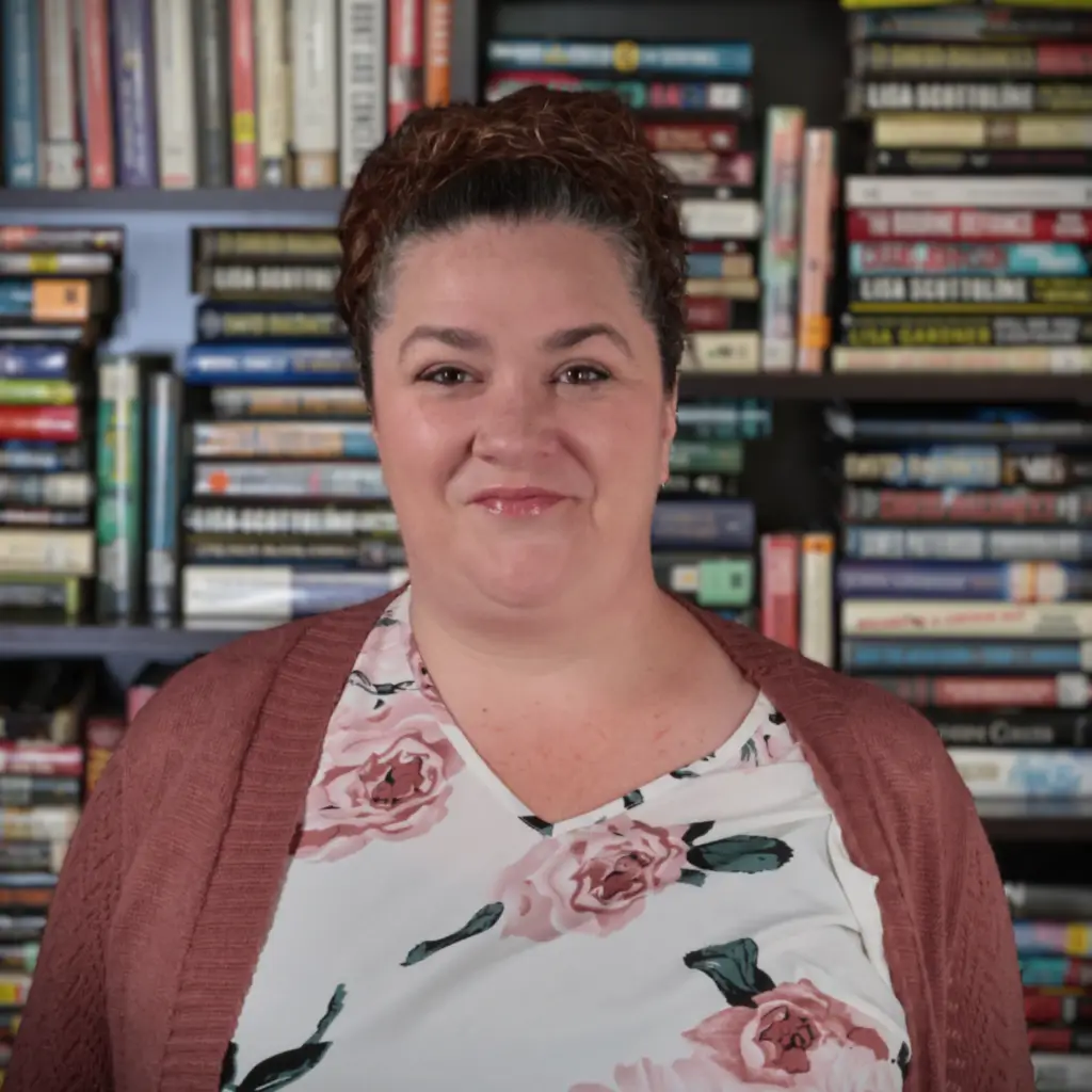 Close-up portrait of a woman in a floral blouse and maroon cardigan, standing in front of blurred bookshelves filled with colorful books
