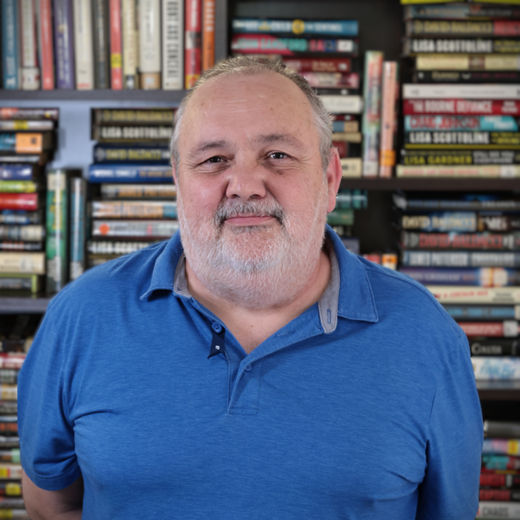 Close-up of an older man with gray beard smiling, wearing a blue shirt, in front of a blurred bookshelf.
